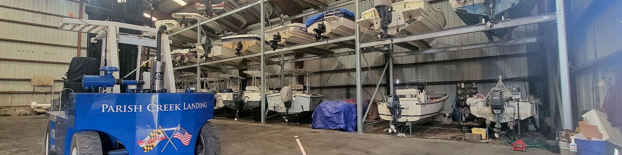 A boatyard hangar with a blue &ldquo;PARISH CREEK LANDING&rdquo; trailer and stored small boats and equipment inside a warehouse.