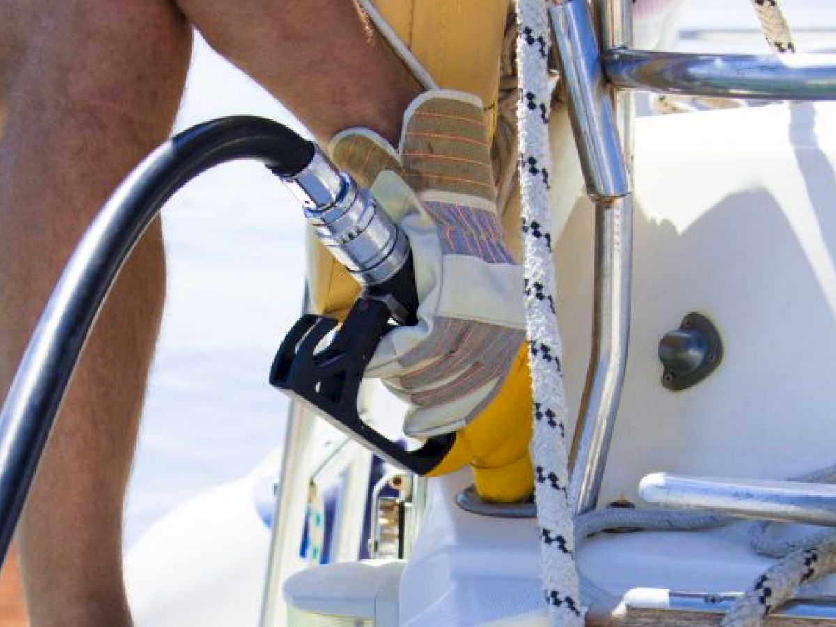 A person is refueling a boat, holding a fuel nozzle next to the fuel intake on the vessel, with some ropes visible.