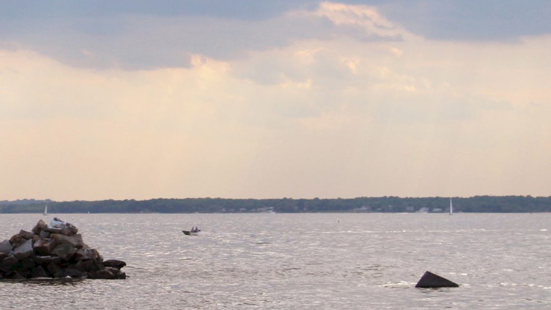 An offshore lighthouse with a red roof stands on stilts, surrounded by rocks in a body of water, with a boat in the distance and a cloudy sky.