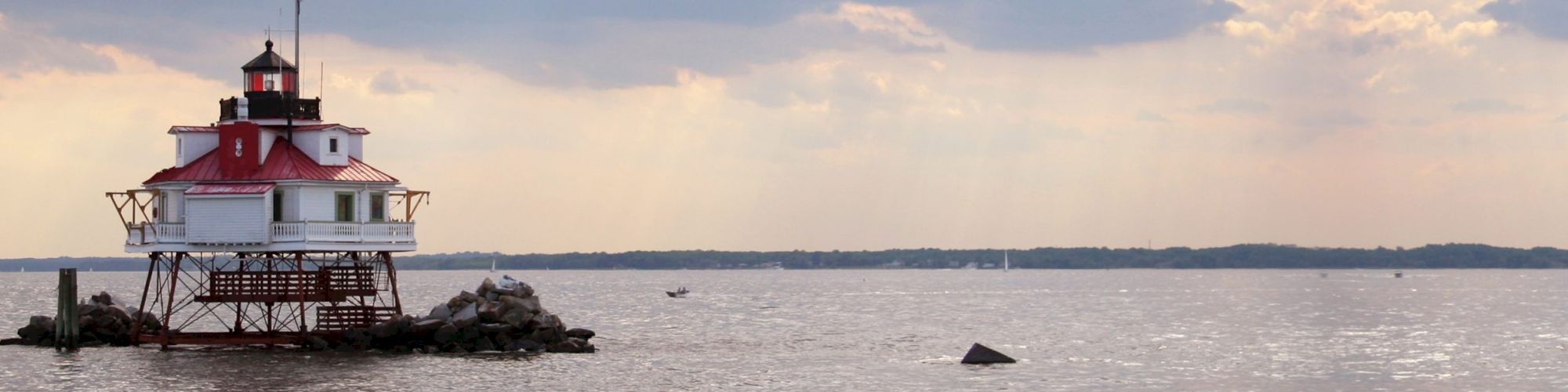 An offshore lighthouse with a red roof stands on stilts, surrounded by rocks in a body of water, with a boat in the distance and a cloudy sky.