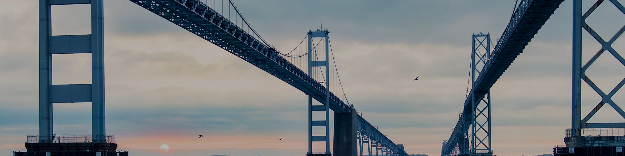 The image shows two parallel bridges over a body of water with a cloudy sky and the sun setting on the horizon.