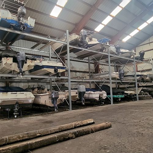 The image shows an indoor storage facility with several boats stacked on metal racks on the left side and a spacious open floor area.
