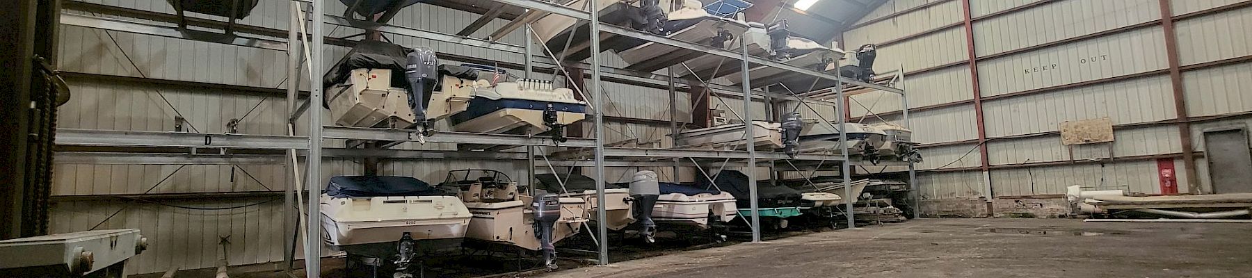 Inside an industrial hangar, several small boats with outboard motors sit on metal racks and platforms under high lights.