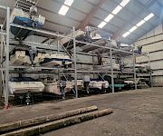 Inside an industrial hangar, several small boats with outboard motors sit on metal racks and platforms under high lights.