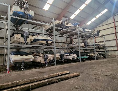 Inside an industrial hangar, several small boats with outboard motors sit on metal racks and platforms under high lights.