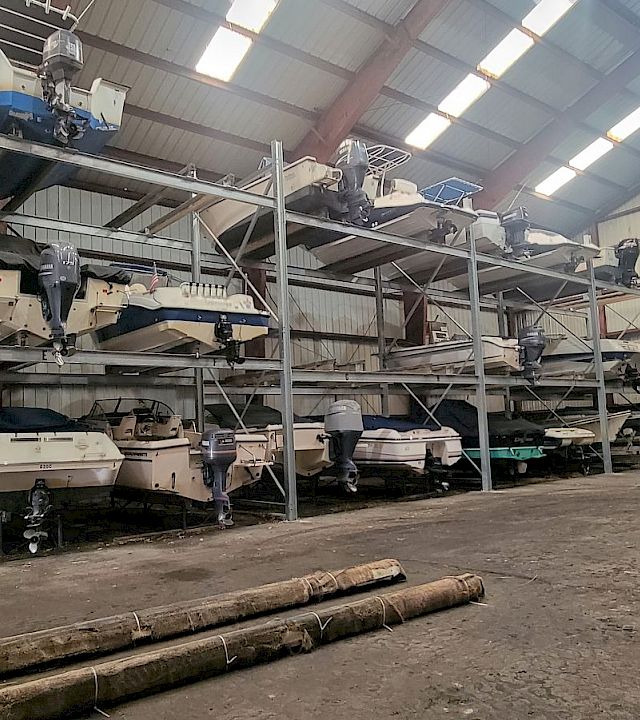 Inside an industrial hangar, several small boats with outboard motors sit on metal racks and platforms under high lights.