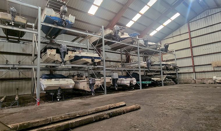 Inside an industrial hangar, several small boats with outboard motors sit on metal racks and platforms under high lights.