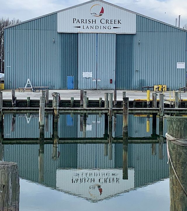 A “Parish Creek Landing” warehouse sits by a marina, with docks, boats, and calm water reflecting the building.