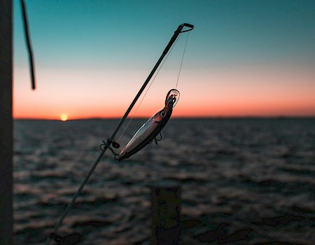 A fishing rod and lure fly above the beach at sunset, with the ocean horizon glowing behind.