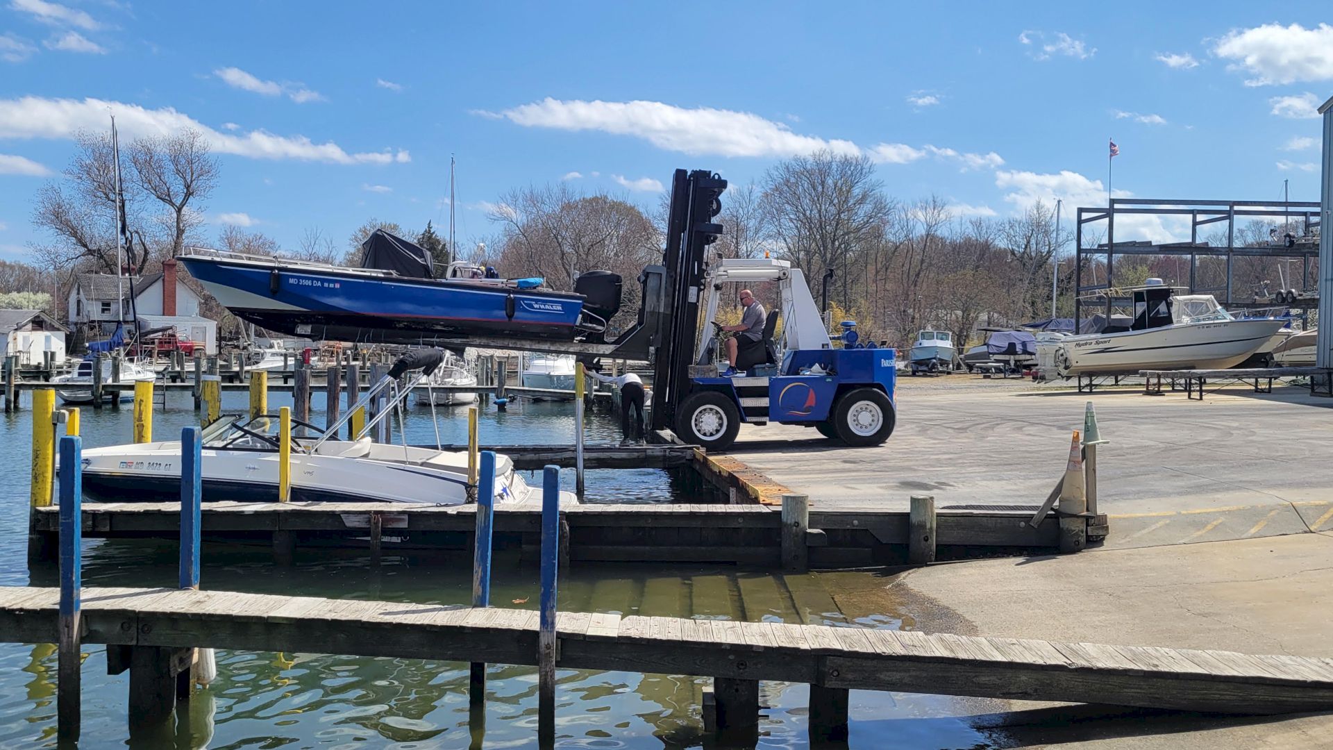A forklift lifts a speedboat on a dock beside other boats at a marina on a sunny day.