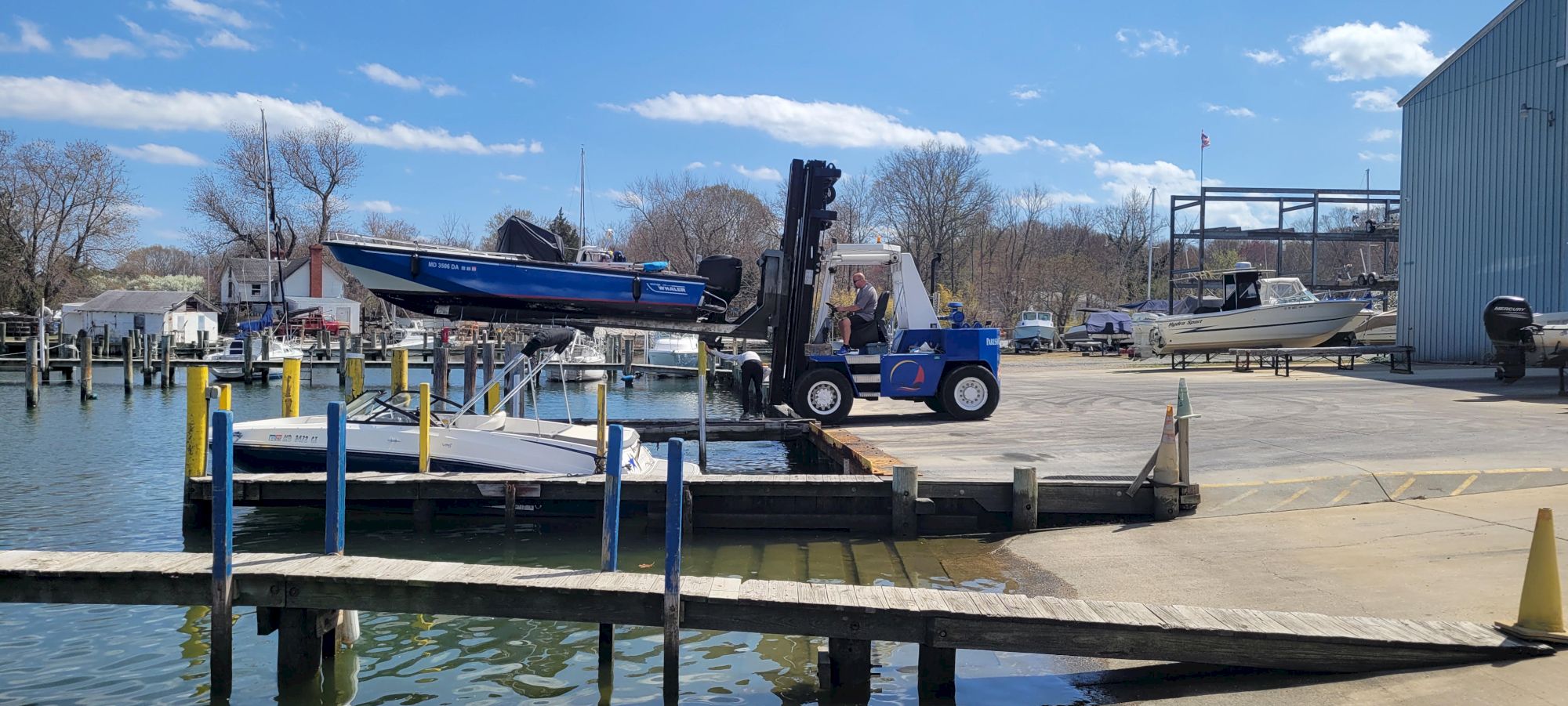 A forklift lifts a speedboat on a dock beside other boats at a marina on a sunny day.
