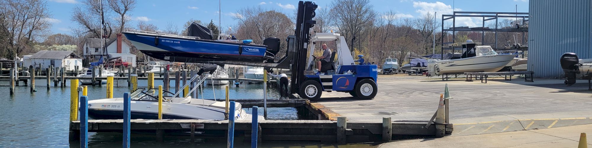 A marina dock with a forklift crane lifting a blue speedboat onto a trailer, while other boats sit in the background.
