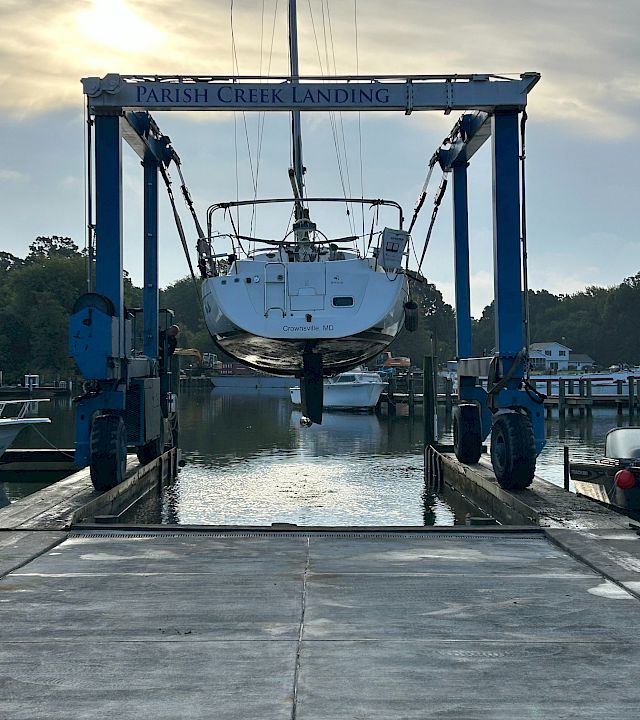 A boat named “Crownsville, MD” is lifted by a crane at Parish Creek Landing in a marina under cloudy skies.