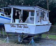 A small motorboat named “Miss Betty Jane” sits on a trailer with a canopy and dock gear, parked by trees.