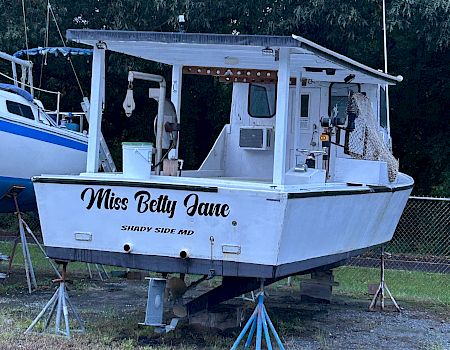 A small motorboat named “Miss Betty Jane” sits on a trailer with a canopy and dock gear, parked by trees.