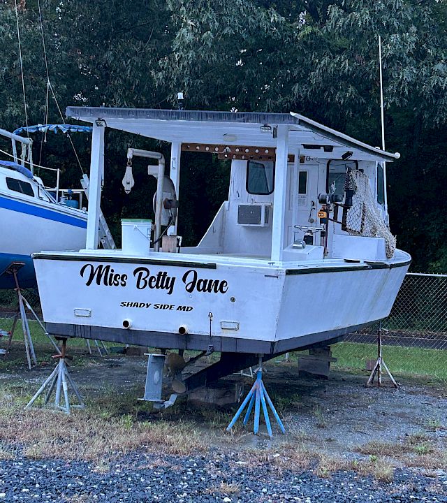 A small motorboat named “Miss Betty Jane” sits on a trailer with a canopy and dock gear, parked by trees.