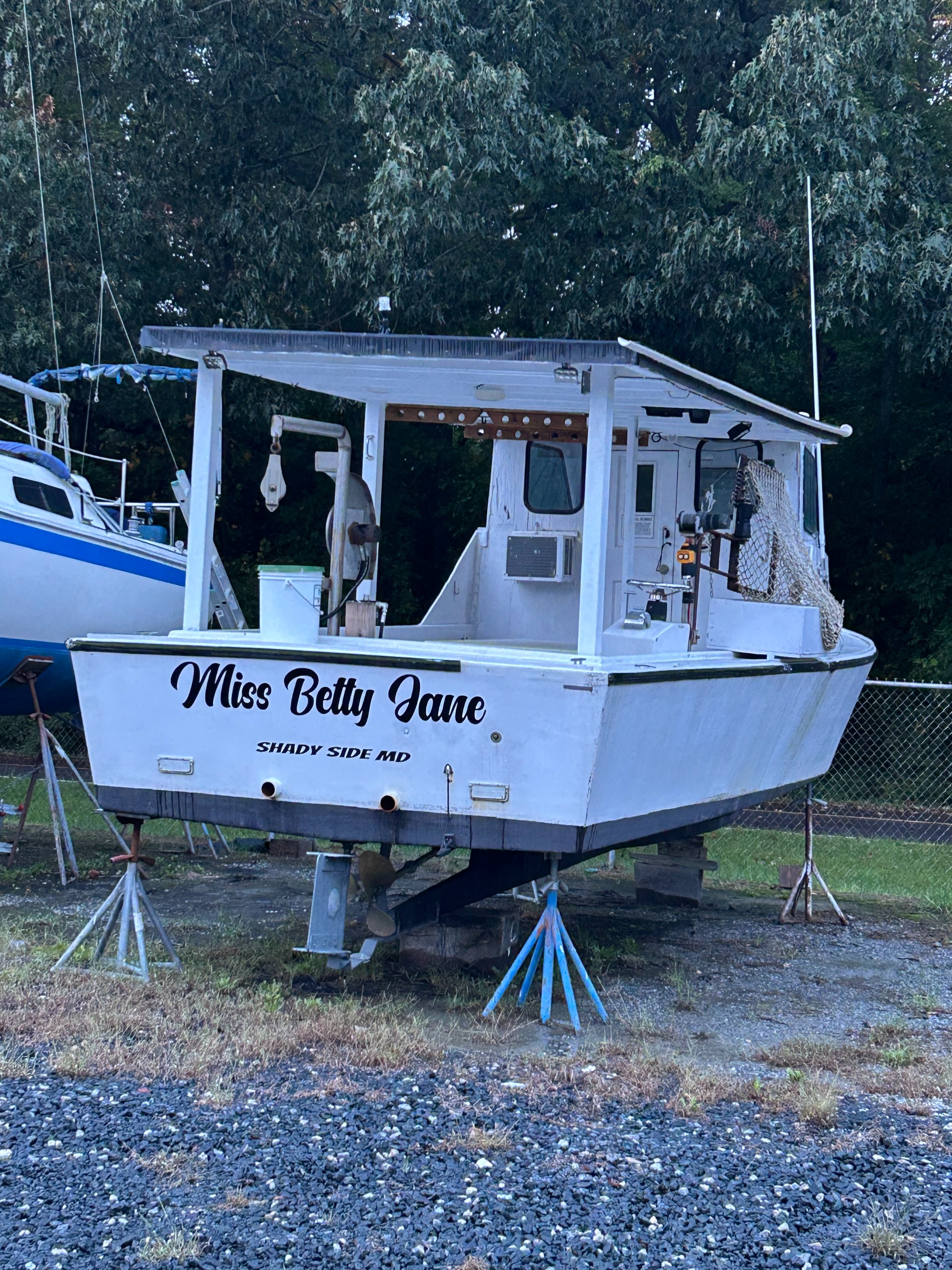 A small motorboat named “Miss Betty Jane” sits on a trailer with a canopy and dock gear, parked by trees.