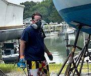 A masked person on a dock, wearing a life vest, posing by a river with boats and a forested shoreline behind them.