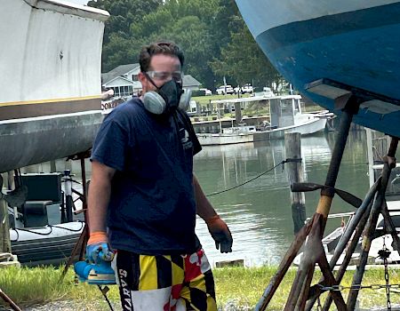 A masked person on a dock, wearing a life vest, posing by a river with boats and a forested shoreline behind them.