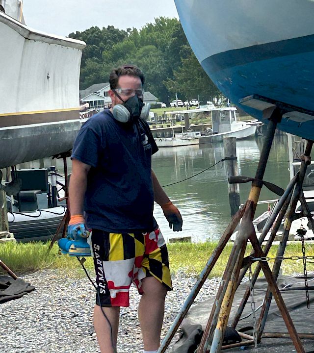 A masked person on a dock, wearing a life vest, posing by a river with boats and a forested shoreline behind them.