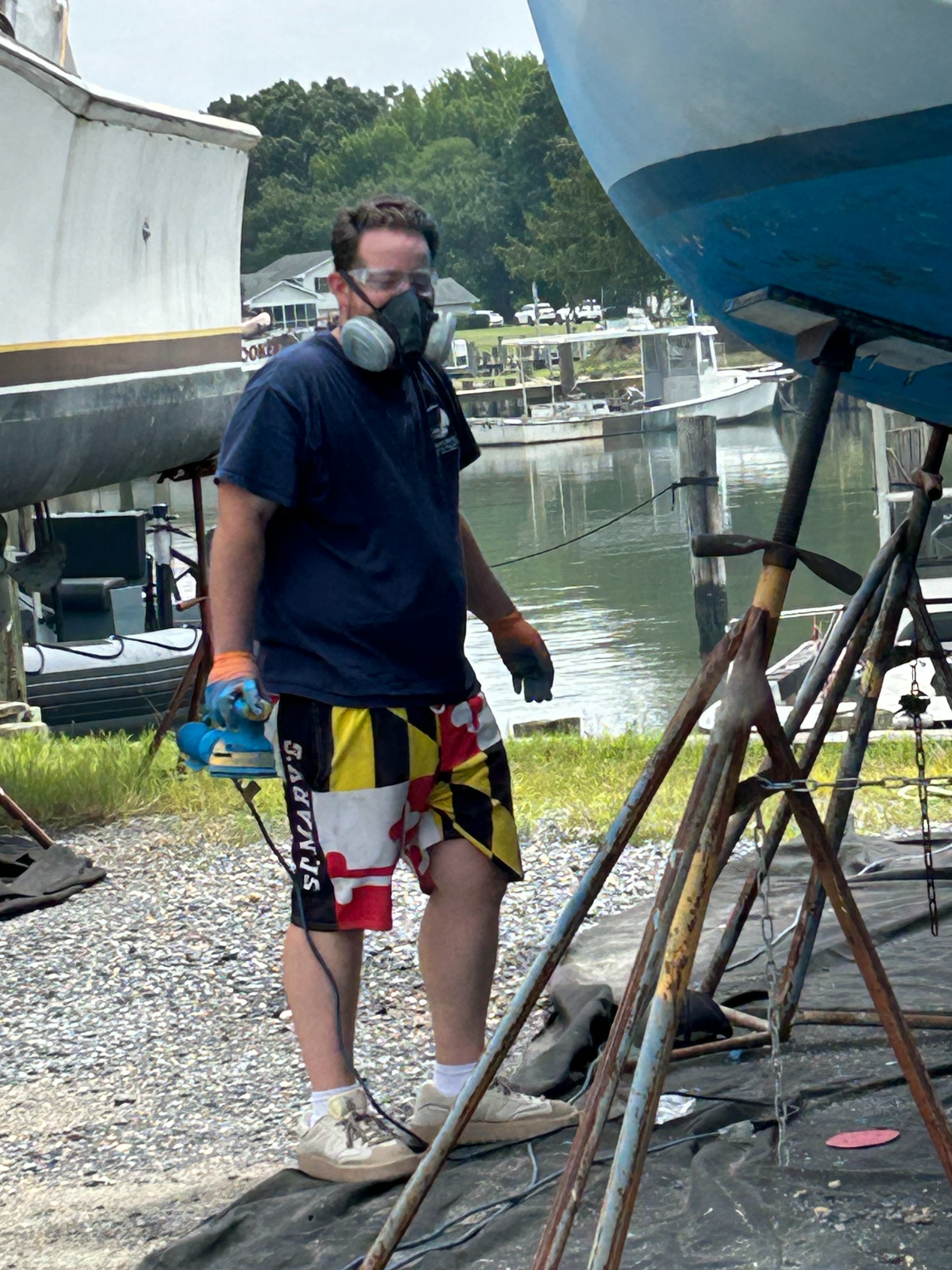 A masked person on a dock, wearing a life vest, posing by a river with boats and a forested shoreline behind them.