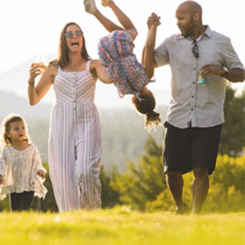 A family is enjoying a sunny day outdoors. The parents are playfully swinging a child, while another child walks alongside them, smiling.