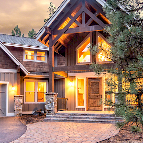 A modern, two-story house with a large driveway, multiple windows, a warm exterior, and surrounded by trees under a dusk sky.