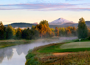 A serene landscape featuring a golf course, a calm river, trees, and a snow-capped mountain under a partly cloudy sky during sunrise or sunset.