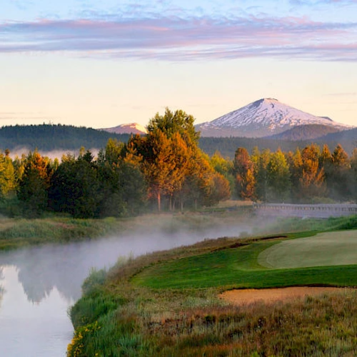 A serene landscape featuring a golf course, a calm river, trees, and a snow-capped mountain under a partly cloudy sky during sunrise or sunset.
