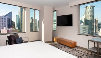 A modern hotel room features a bed, armchair, TV, console, and large windows offering a city skyline view, including several high-rise buildings.