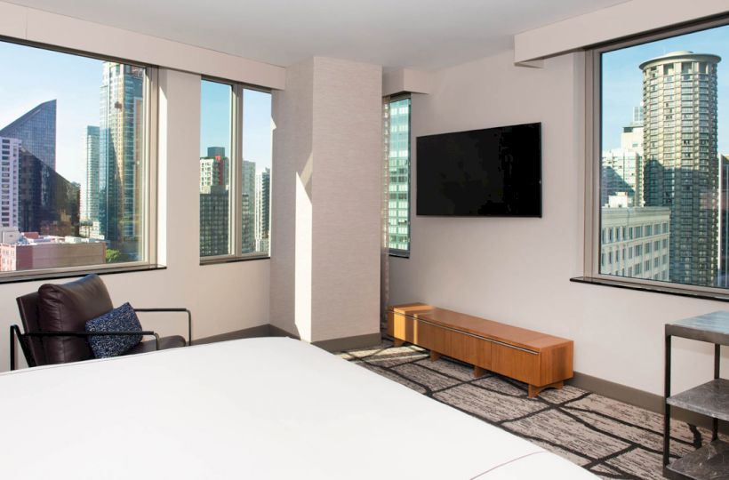 A modern hotel room features a bed, armchair, TV, console, and large windows offering a city skyline view, including several high-rise buildings.