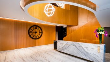 A modern lobby with wooden walls, a marble reception desk, a large clock, spiral architectural elements, spherical lights, and a vase with flowers.