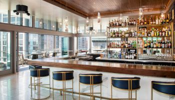 The image shows a modern bar area with a long wooden bar, high chairs, shelves stocked with bottles, and large windows providing a city view.