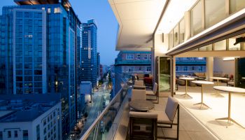 The image shows a modern rooftop terrace with comfortable seating and tables, overlooking a cityscape with tall buildings at dusk.