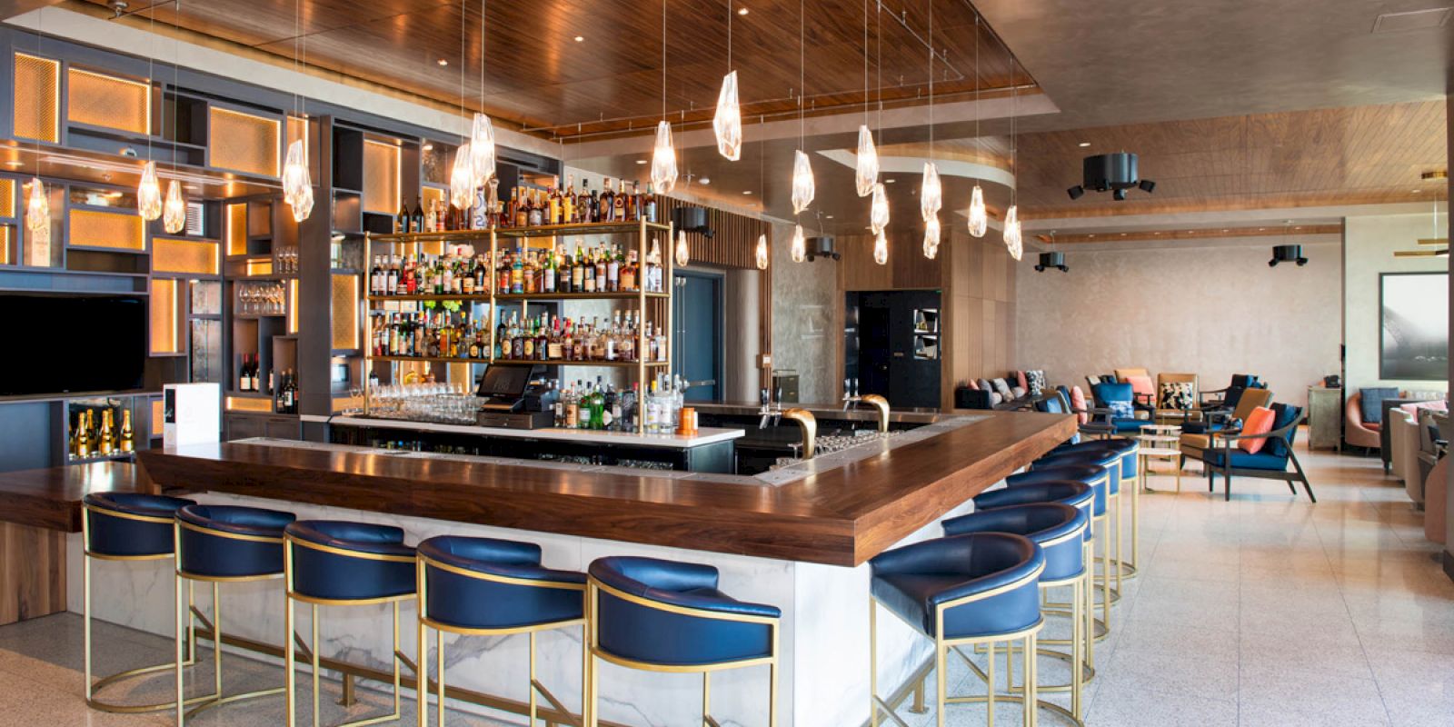 This image shows a modern bar with a wooden counter, stylish chairs, hanging lights, and a well-stocked liquor shelf. It has a spacious, cozy lounge area.