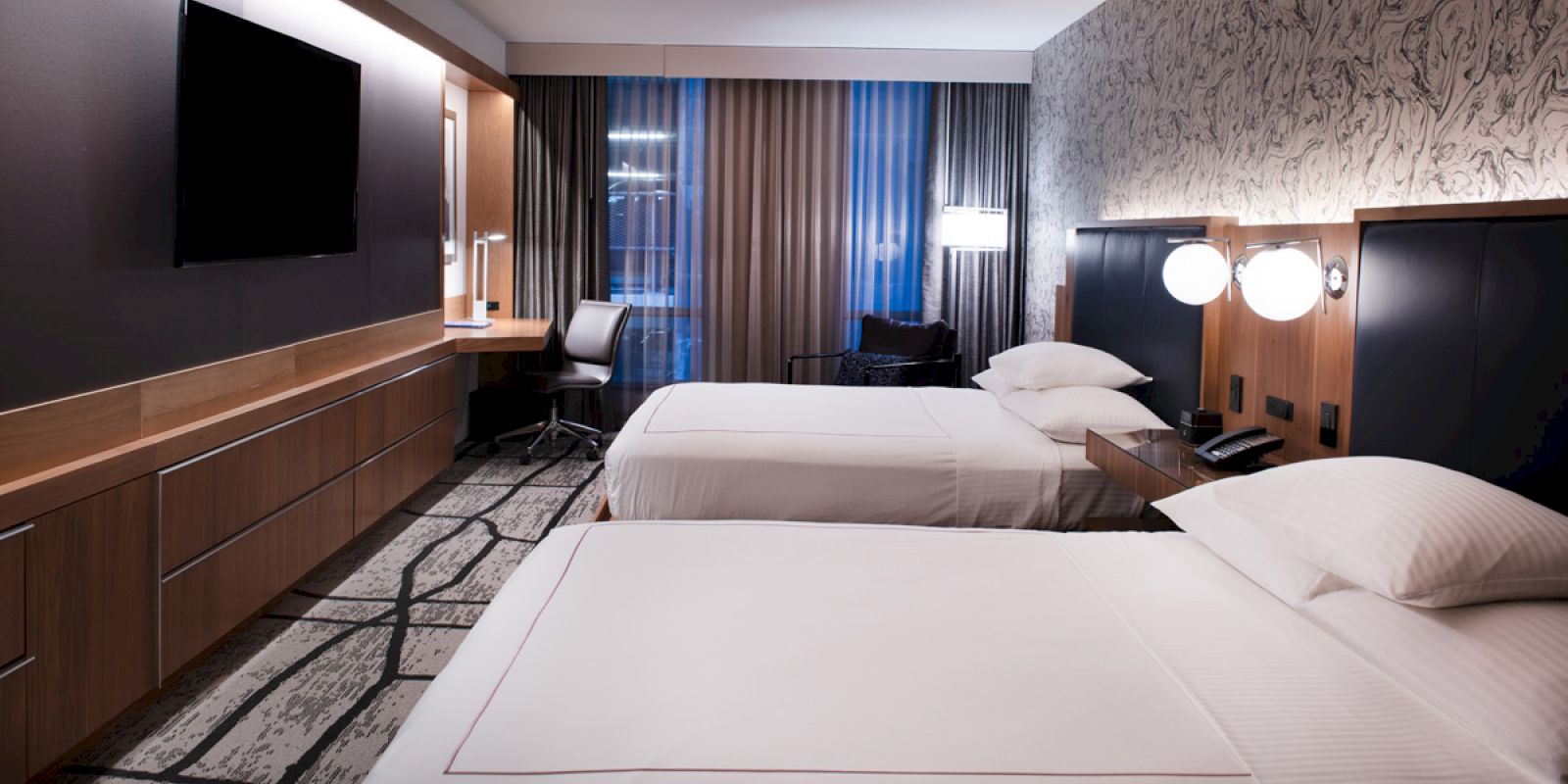 A modern hotel room with two beds, flat-screen TV, desk with chair, and contemporary decor featuring a patterned wall and ambient lighting.
