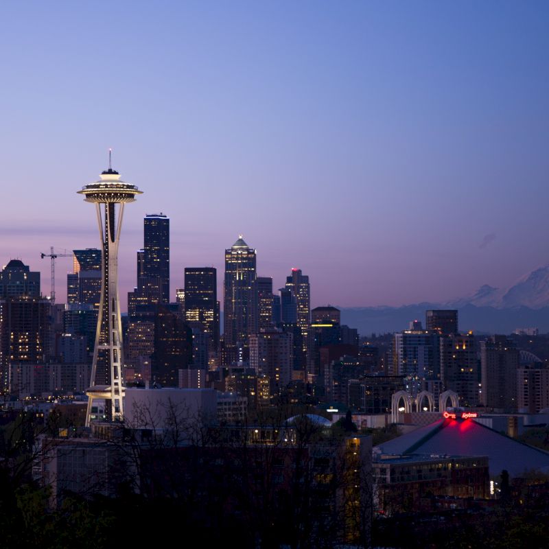 The image shows the Seattle skyline at dusk, featuring the Space Needle prominently. The cityscape is illuminated with Mount Rainier visible in the background.