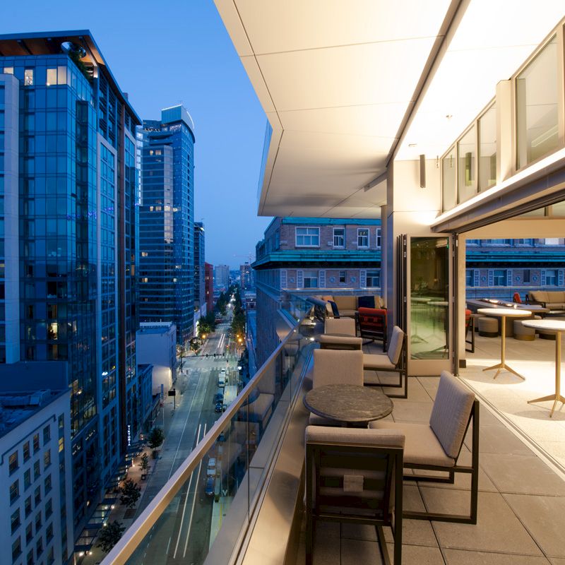 The image shows a modern high-rise cityscape at dusk with a view from a luxurious balcony, featuring seating and lighting, overlooking buildings.