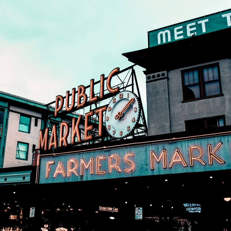A neon sign that reads "PUBLIC MARKET" and "FARMERS MARKET" with a clock is mounted on a building. Nearby signs include "ARCADE SHOPS" and "MEET THE."