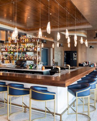 A modern bar with wooden countertops, stylish barstools, and a well-stocked liquor shelf under hanging pendant lights in a spacious setting.