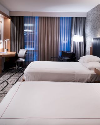 The image shows a modern hotel room with two neatly made beds, a flat-screen TV, a work desk, and a stylish, patterned wall.
