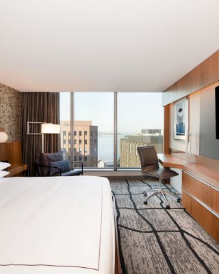 A modern hotel room features a large bed, a wall-mounted TV, a desk with a chair, and a window with a city view in the background.