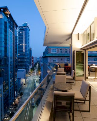 The image shows a modern rooftop terrace with seating and tables, overlooking a city street lined with tall buildings at dusk.
