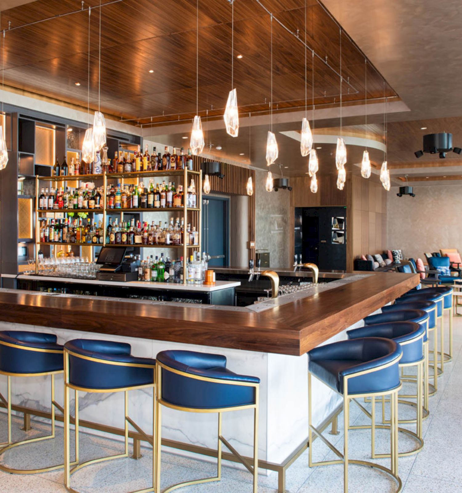 A modern, upscale bar with a long wooden counter, blue stools, a well-stocked liquor shelf, pendant lights, and a seating area with tables and chairs.