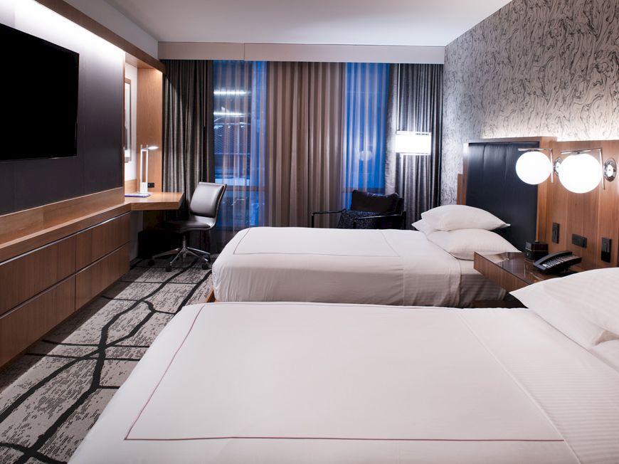 The image shows a modern hotel room with two neatly made beds, a TV, desk, chair, and stylish lighting, featuring contemporary decor.