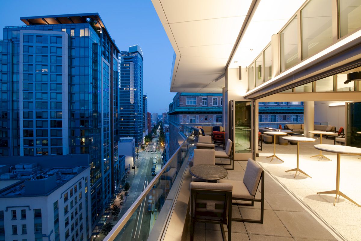 A modern balcony overlooking a cityscape at dusk, with tables and chairs arranged around it, and skyscrapers with lights in the background.