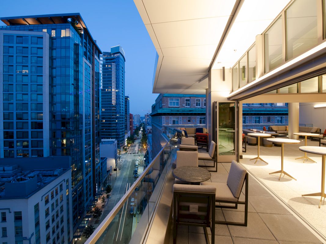 A modern balcony overlooking a cityscape at dusk, with tables and chairs arranged around it, and skyscrapers with lights in the background.