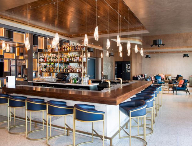 A modern bar with a wooden countertop, blue chairs, numerous light fixtures, and a well-stocked shelf of bottles. There is seating in the background.