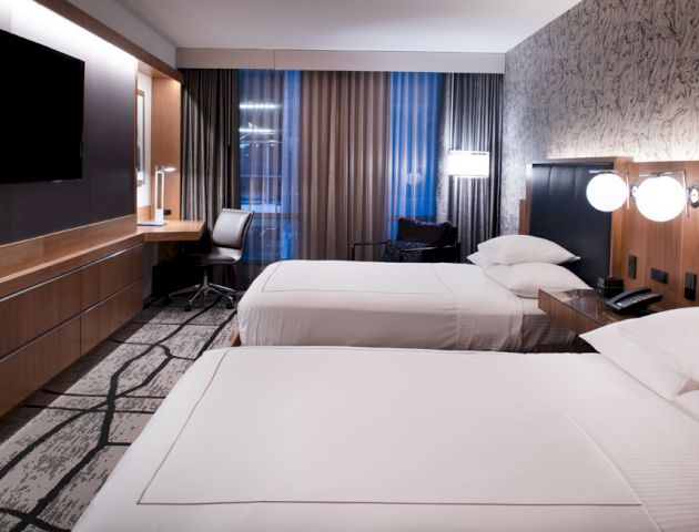 A modern hotel room with two neatly made beds, a flat-screen TV, a desk with a chair, large windows with curtains, and a patterned wall.