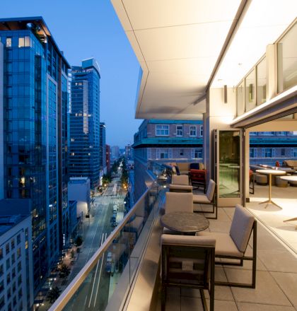 A modern cityscape at dusk with high-rise buildings. The view is from a balcony featuring tables and chairs with a partially enclosed space.